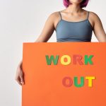 Woman with short hair holding a vibrant 'Work Out' sign on a white background.